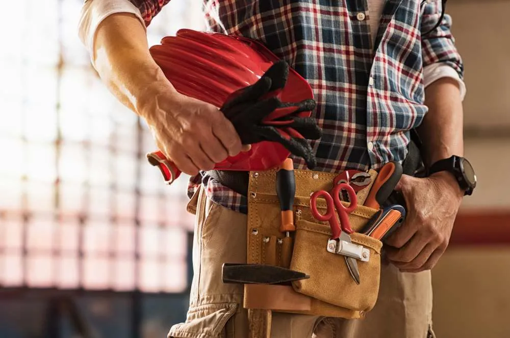 Handyman with a loaded tool belt, holding a hard hat and work gloves
